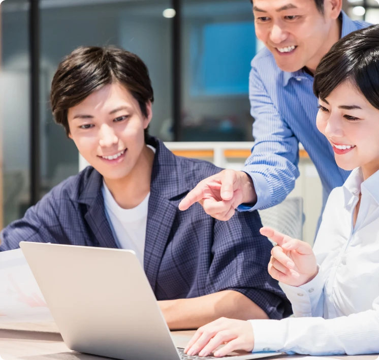 先輩社員に教わりながら、笑顔でパソコン画面を覗き込む若い男女のオフィス風景