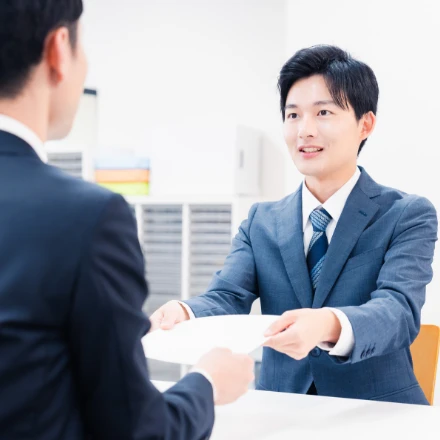 オフィスの受付や対面ブースで、笑顔で丁寧に書類を手渡すスーツ姿の若い男性スタッフ
