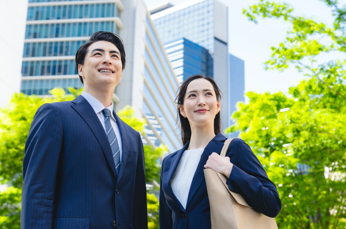 高層ビル街をバックに、笑顔で未来を見つめるように上を見上げるスーツ姿の若い男女のビジネスパーソン