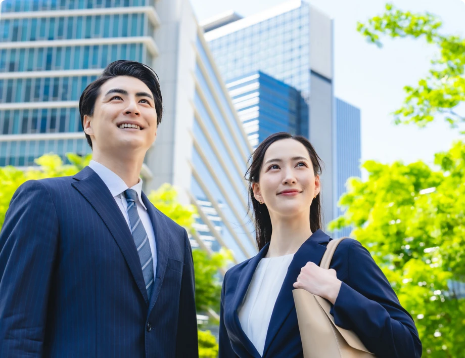 高層ビル街をバックに、笑顔で未来を見つめるように上を見上げるスーツ姿の若い男女のビジネスパーソン