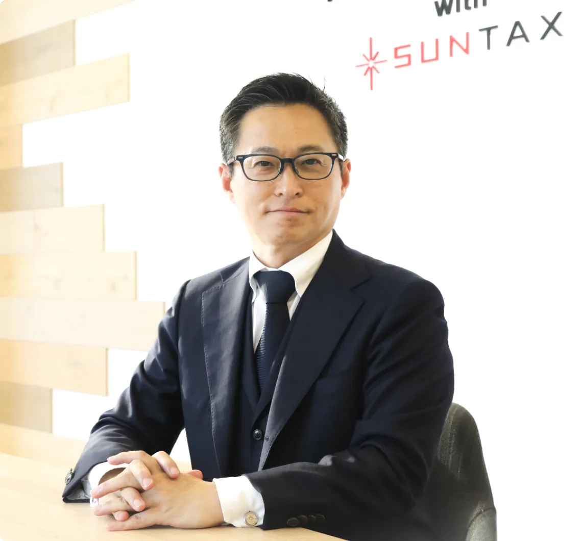 SUNTAXのロゴの前で、テーブルに手を置き真摯な表情で正面を見つめるスーツ姿の代表者
