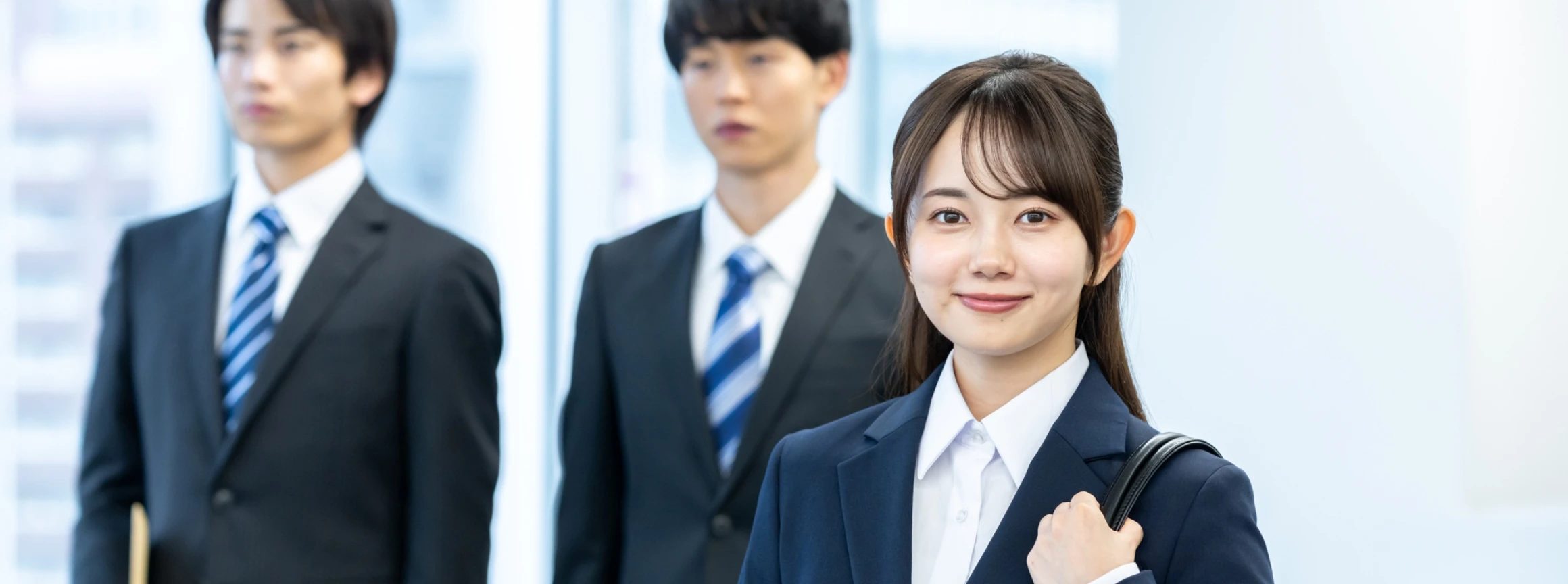 オフィスカジュアルの男性社員を背景に、スーツ姿で前を向いて自信に満ちた笑顔浮かべる女性社員
