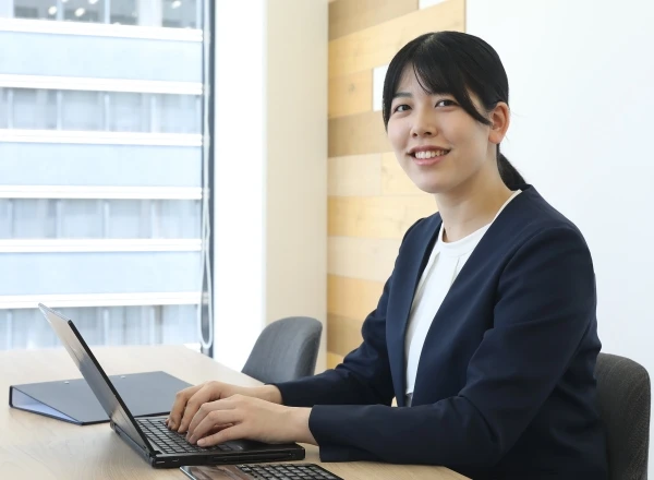 オフィスのデスクでノートパソコンを操作しながら、笑顔でカメラを見つめる女性社員