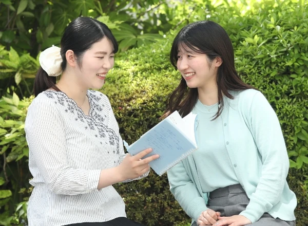 緑豊かな屋外のベンチで、ノートを広げて楽しそうに話す2人の女性社員