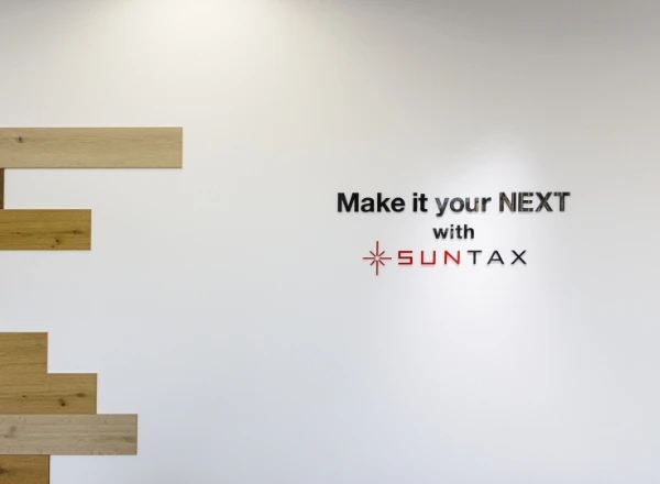 「Make it your NEXT with SUNTAX」のコーポレートロゴが書かれたオフィスの白い壁