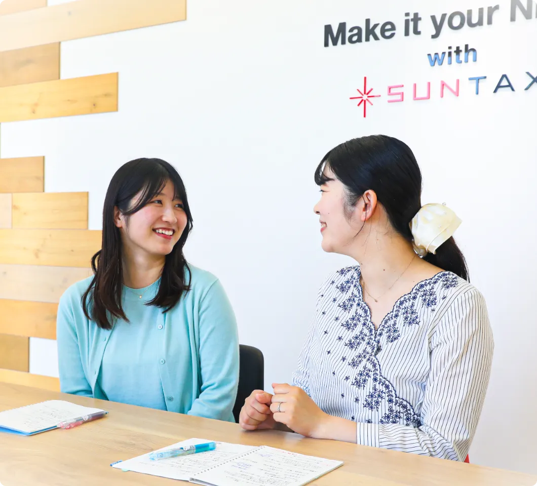 SUNTAXのロゴの前で、ノートを広げて笑顔で打ち合わせをする2人の女性社員