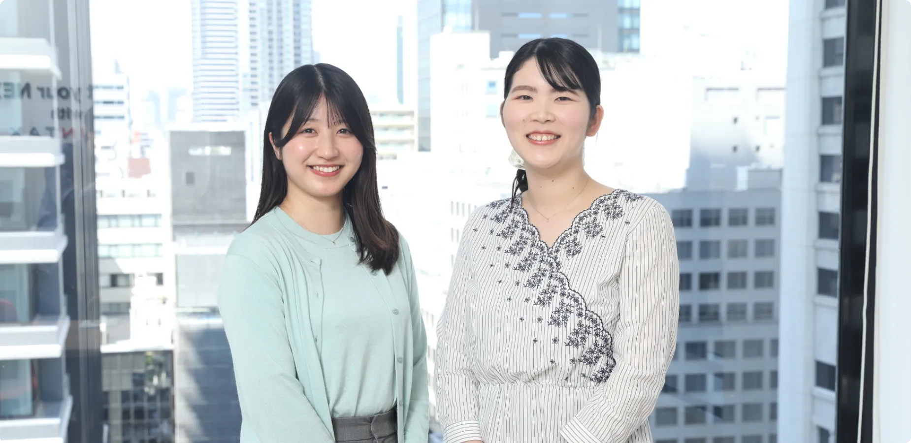オフィスの窓際で街並みをバックに、並んで笑顔を見せる2人の女性社員