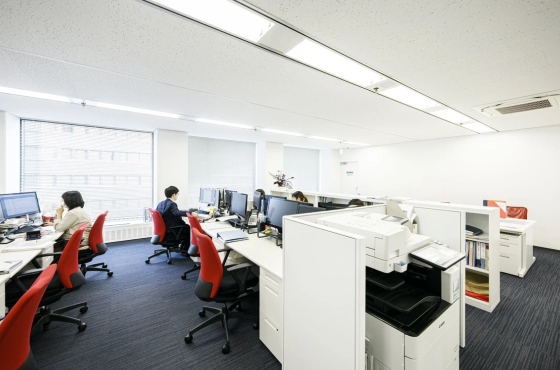 白いデスクに赤いオフィスチェアが映える、社員がパソコンに向かって業務を行う執務スペースの全景