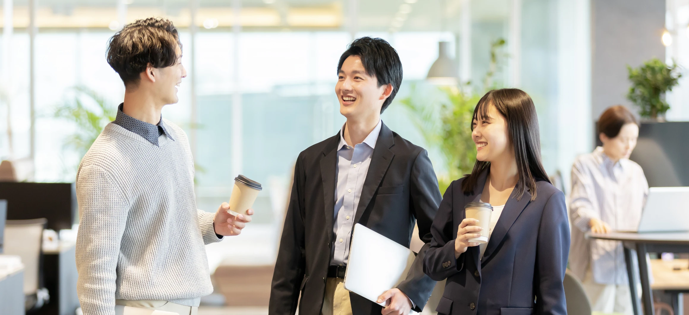 明るいオフィスでノートパソコンやコーヒーを手に、笑顔で談笑しながら歩く若い男女の会社員たち