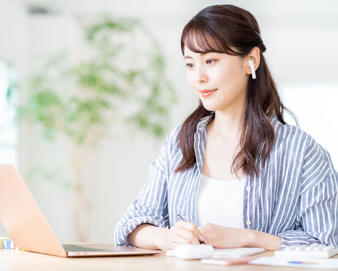 イヤホンを着用し、ノートパソコンを開いてメモを取りながらオンライン研修を受ける女性社員