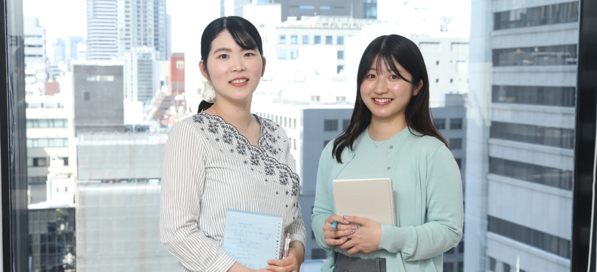 オフィスの大きな窓を背景に、ノートを手にして笑顔で並ぶ2人の女性社員