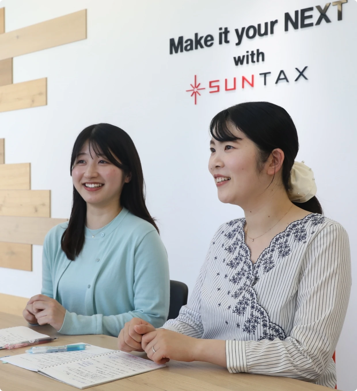 SUNTAXのロゴがデザインされた会議室で、ノートを前に笑顔で話を聞く2人の女性社員