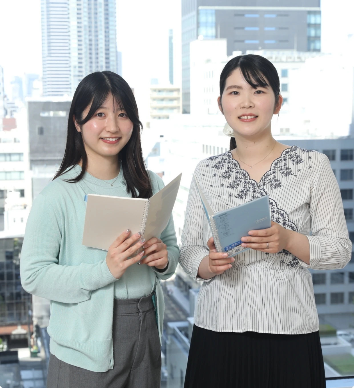都会の景色をバックに、それぞれノートを開いて自信に満ちた表情で佇む2人の女性社員