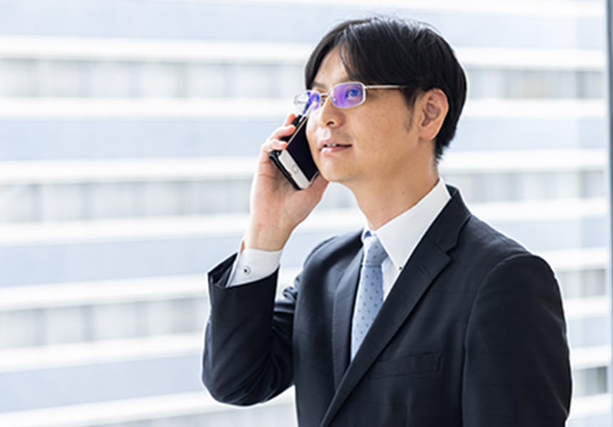 スーツ姿の男性が窓際でスマートフォンを耳にあてて電話している様子